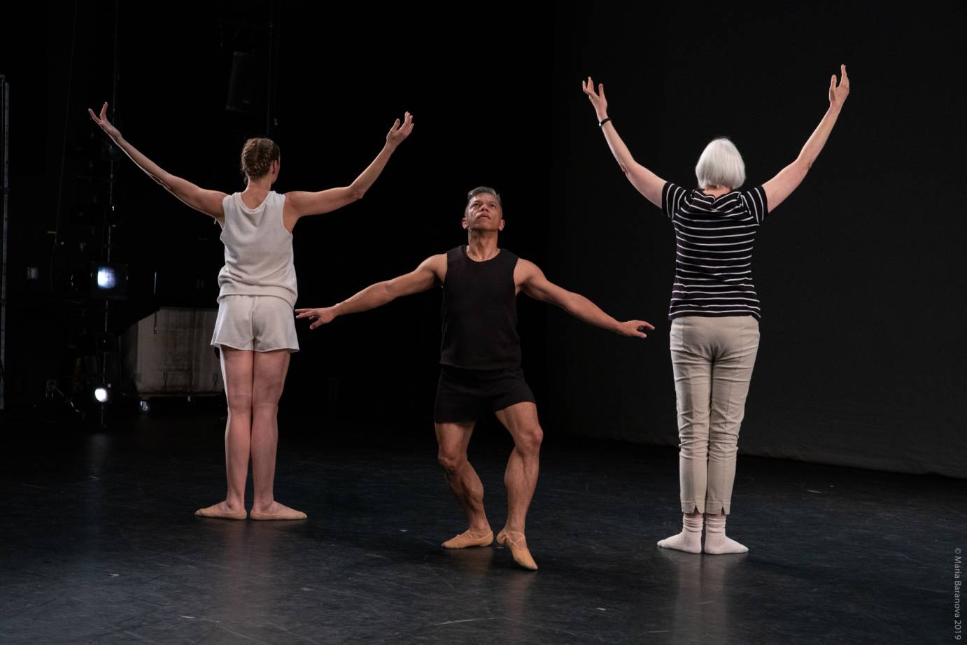 Two women extend their arms in high Vs, backs to audience, while one man extends his arms in a low V